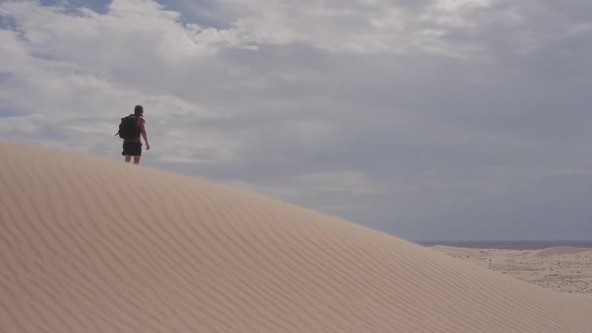 A man wearing a backpack and hat stands on a high sand dune as wind blows sand across the ridge under a cloudy sky