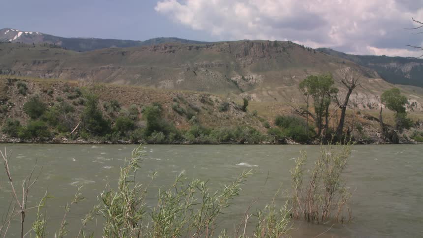 River flowing in front of dry hills with riparian vegetation under a partly cloudy sky