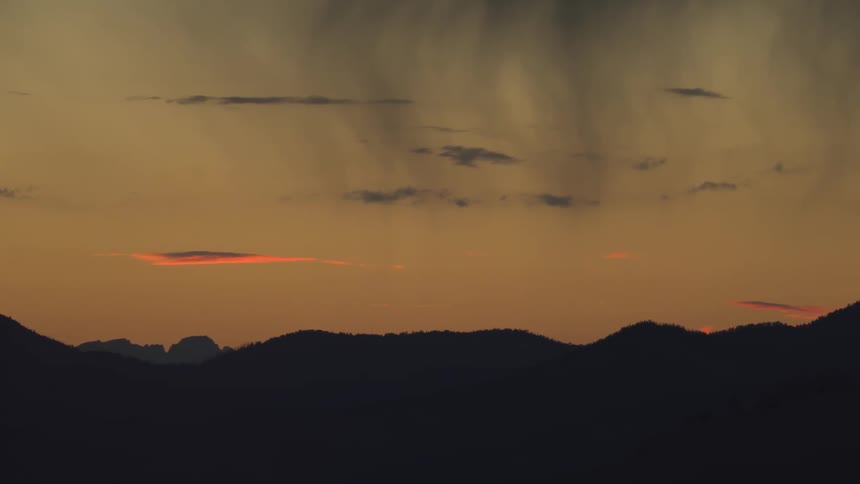 A wide shot shows dark mountain ridges silhouetted against a golden sunset sky where streaks of rain fall from heavy clouds above the horizon