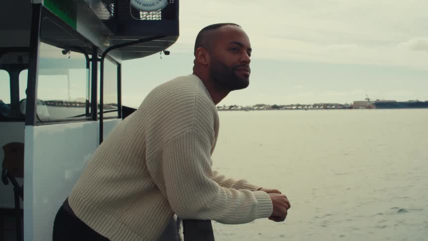 A medium shot shows a man wearing a cream sweater leaning against a railing on a boat as he looks out over the water toward a distant shoreline under a cloudy sky