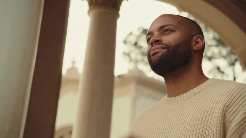 A man in a beige sweater stands near arched stone pillars and smiles as he uses a small point and shoot camera to take a photo of the surrounding architecture during a bright day outdoors