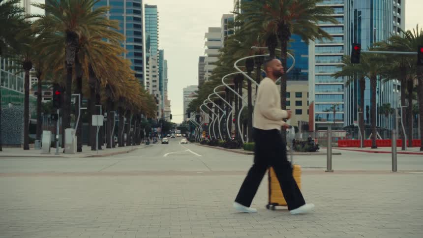 A man walks across a city street pulling a bright yellow suitcase past rows of tall palm trees and modern glass skyscrapers