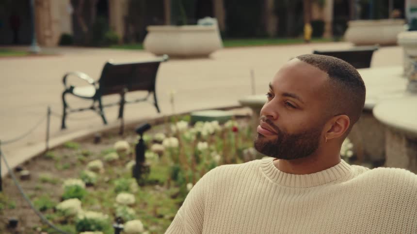 A medium shot shows a man in a cream sweater sitting on a stone ledge outdoors as he looks around a plaza with park benches and flower beds