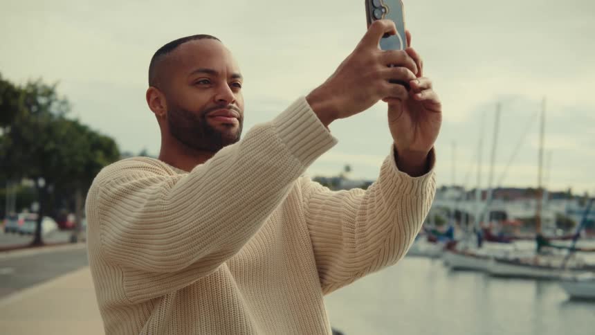 Man stands along the coast with boats in the harbor as he holds up his phone and takes a picture