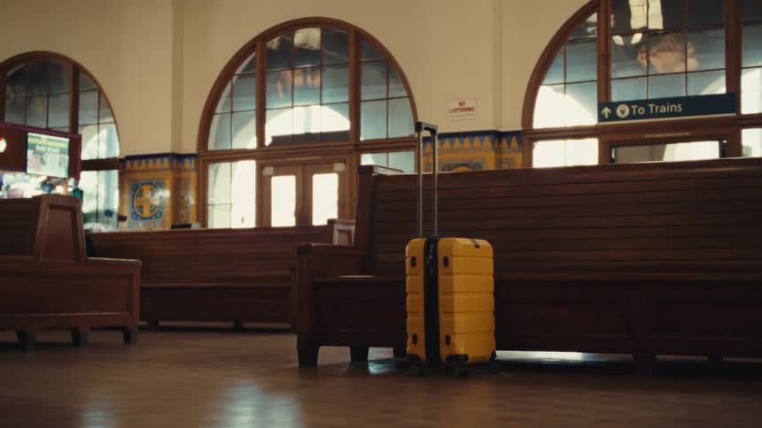 A bright yellow suitcase stands beside a wooden bench inside a quiet train station with large arched windows and a sign pointing to the trains