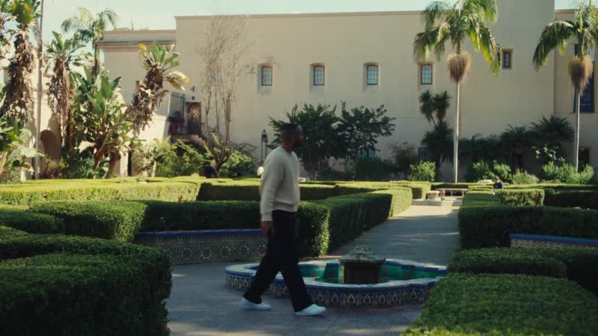 A man walks through a landscaped garden with trimmed hedges and a small fountain in front of a large building on a sunny day