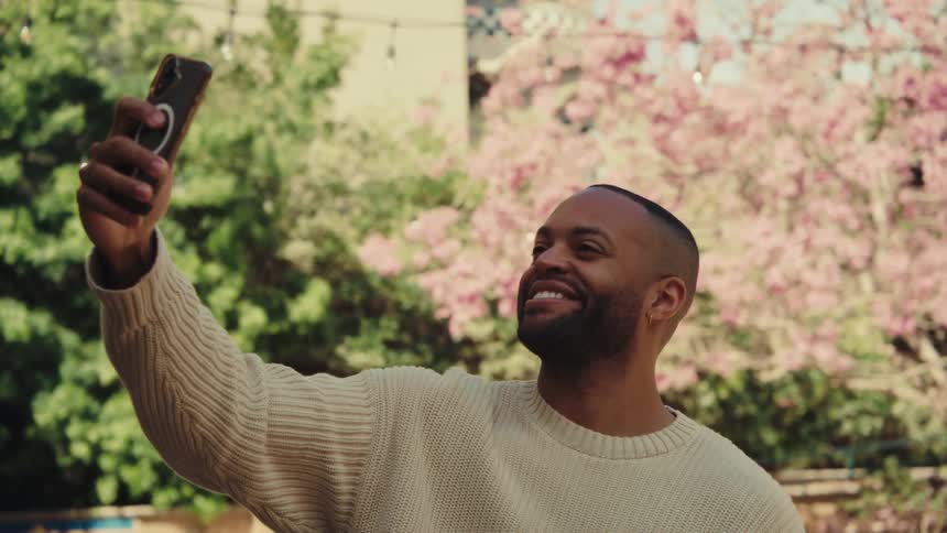 Man smiling while taking a selfie with a smartphone in a garden with blooming trees