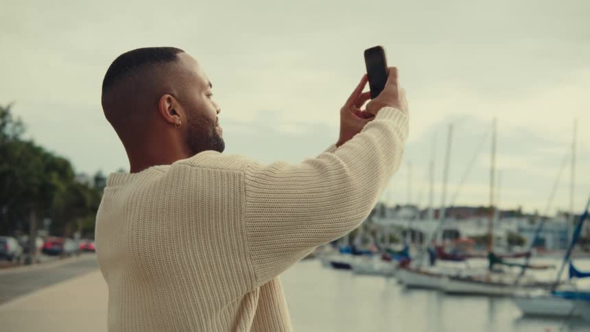A man in a cream sweater holds up his phone to take video footage by a marina filled with boats