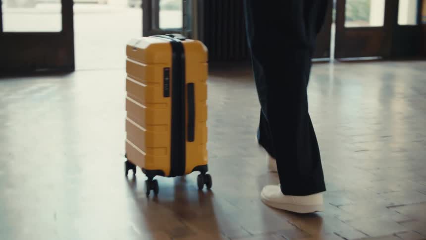 A low angle shot from behind follows a person wearing white sneakers and dark pants as they walk across a tiled floor toward glass doors while pulling a bright yellow suitcase