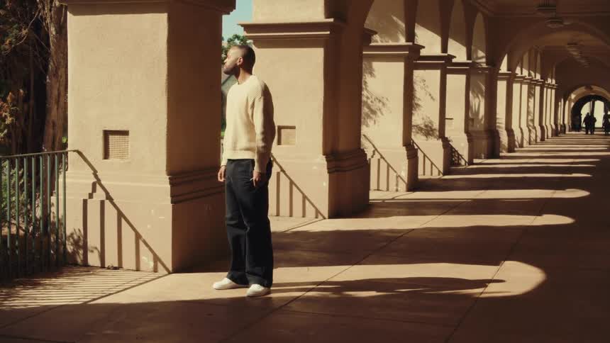 A man stands in a sunlit corridor lined with arches casting long shadows along the walkway with a small group of people down the corridor
