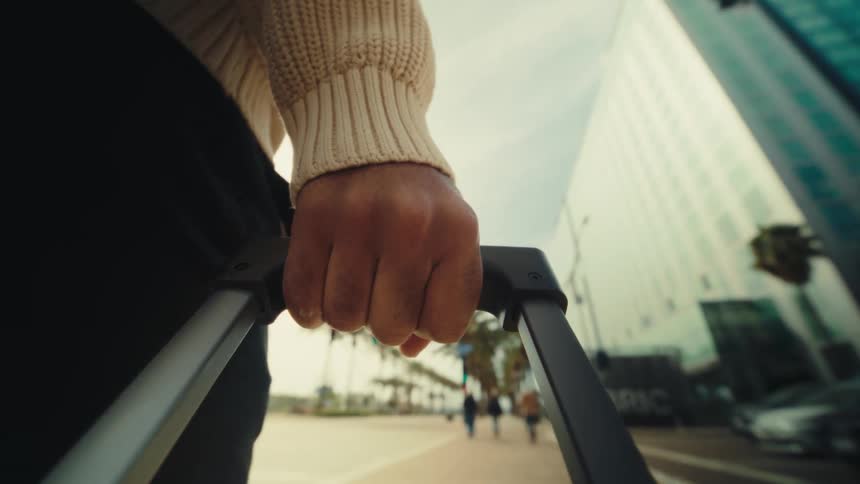 Hand holds the handle of a wheeled suitcase while walking along an urban avenue