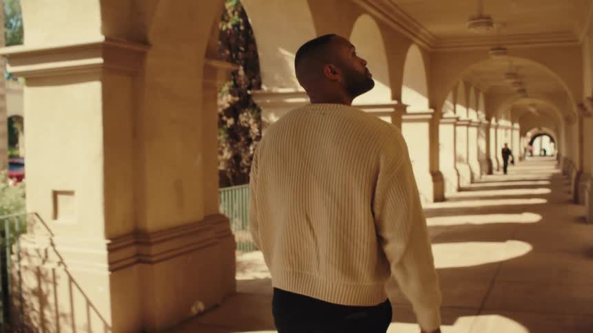 A shot from behind shows a man in a cream sweater as he walks through a long outdoor corridor with stone arches while sunlight creates shadows on the floor