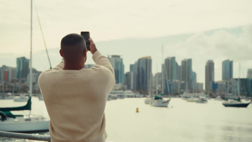Man photographing an urban skyline by a marina with boats