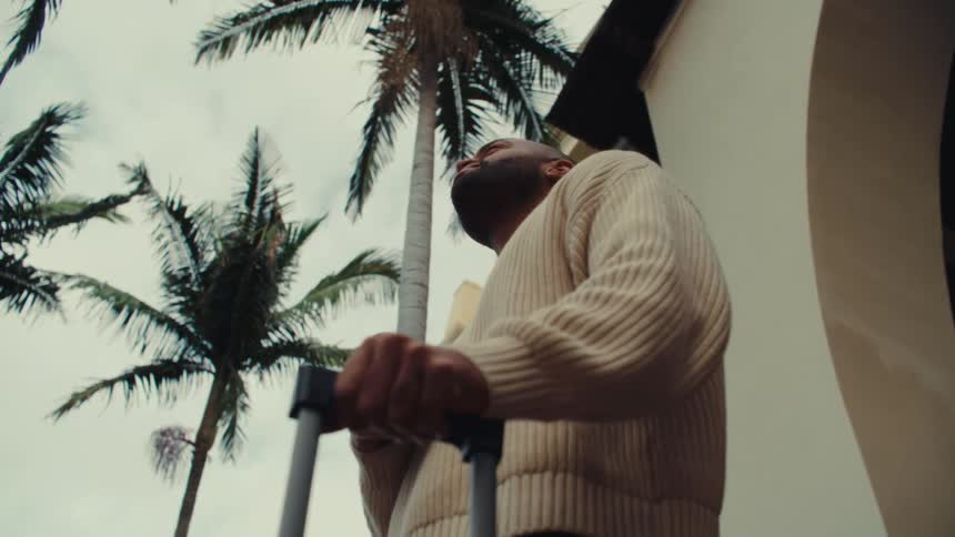 A man steps outside a tropical hotel while carrying his suitcase