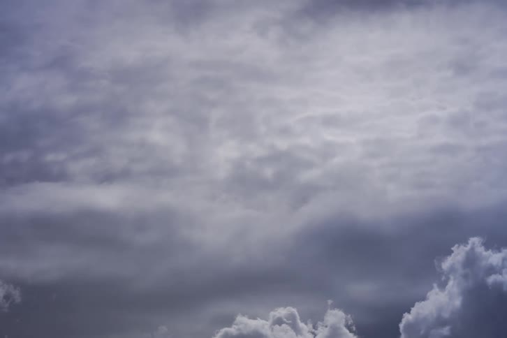 A time lapse view shows thick grey and white clouds moving across the sky and changing shape as they drift under a layer of higher thin clouds during a gloomy day