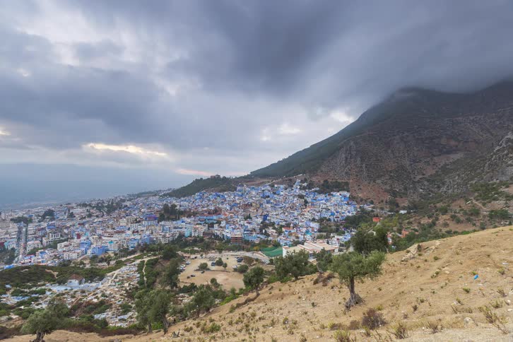 A time lapse view shows a city with blue buildings nestled at the base of a mountain as evening falls and dark clouds move overhead while lights begin to flicker on throughout the streets and homes
