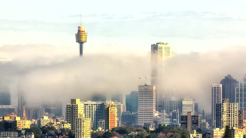 A time lapse view shows a thick layer of white fog rolling through a city skyline and partially obscuring tall office buildings and a prominent observation tower during the morning