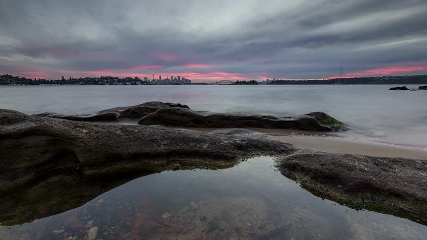 Dark rocks and a small water pool in the foreground with a bay and city skyline at dusk