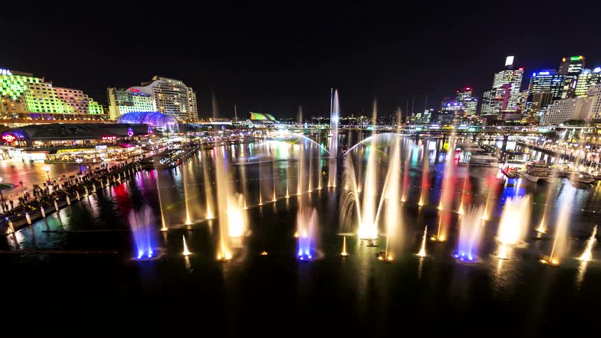 Illuminated fountain with colored water jets in front of a harbor area and lit buildings at night