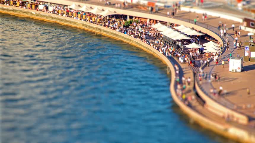 A curved riverside promenade packed with people walking and sitting at an outdoor cafe under white umbrellas next to blue water during a bright sunny afternoon in an urban setting