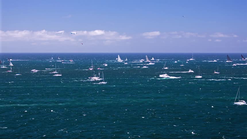 A wide landscape shot of numerous sailboats and yachts navigating a vast choppy blue ocean under a clear sky