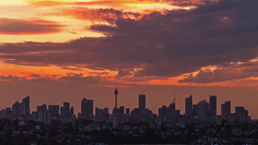 A time lapse view shows a dark city skyline silhouetted against a bright orange and yellow sunset as heavy clouds move across the sky and lights begin to glow in the buildings during the transition to night