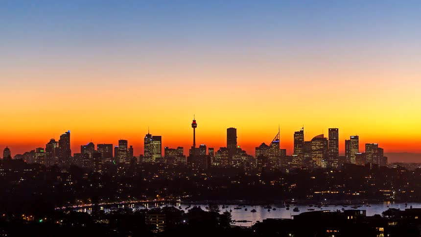 Sun sets over metropolitan buildings and skyscrapers on the sydney australia skyline on the coast as lights come on and traffic moves as night falls