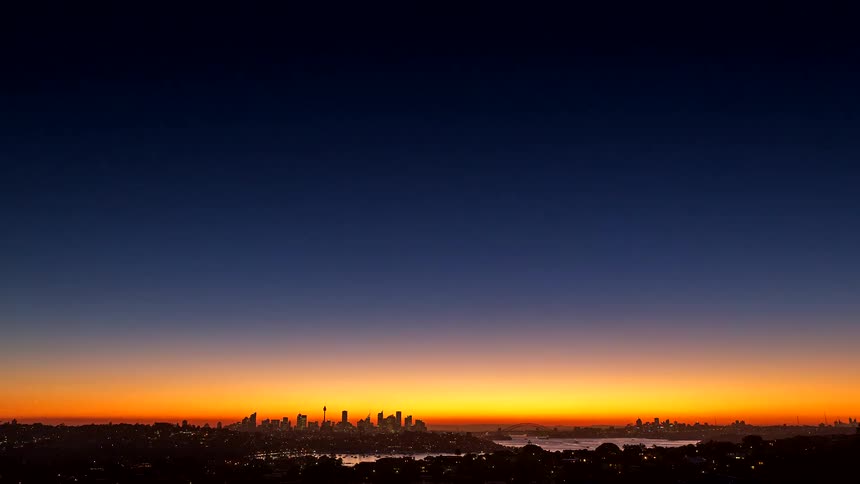A city skyline in the distance with a lake in front under an orange sky as dusk turns into night and the city lights begin to glow