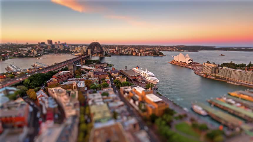 A tilt shift time lapse view shows a high angle of a harbor city with a large bridge and a white opera house as sunlight fades and vibrant city lights illuminate the streets and buildings during a clear evening