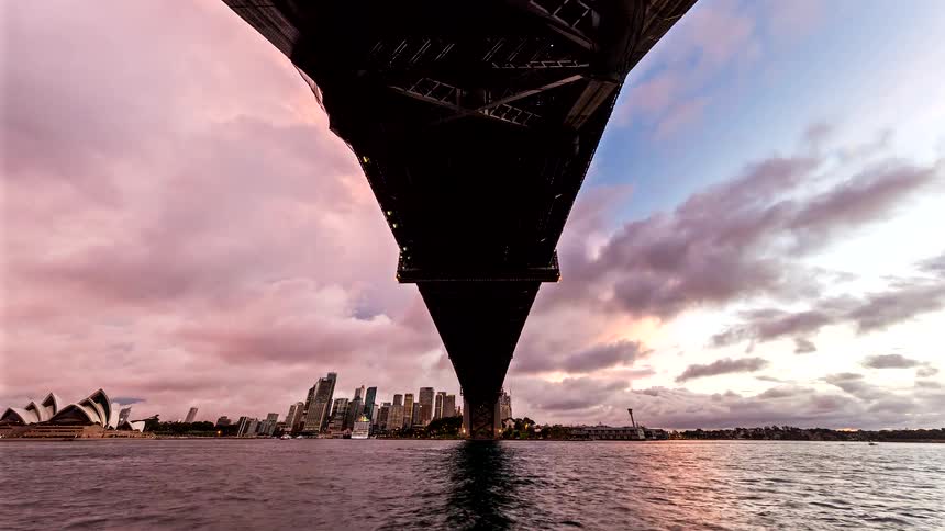 Dramatic view beneath a harbor bridge toward a skyline and waterfront landmark in cloudy dusk light