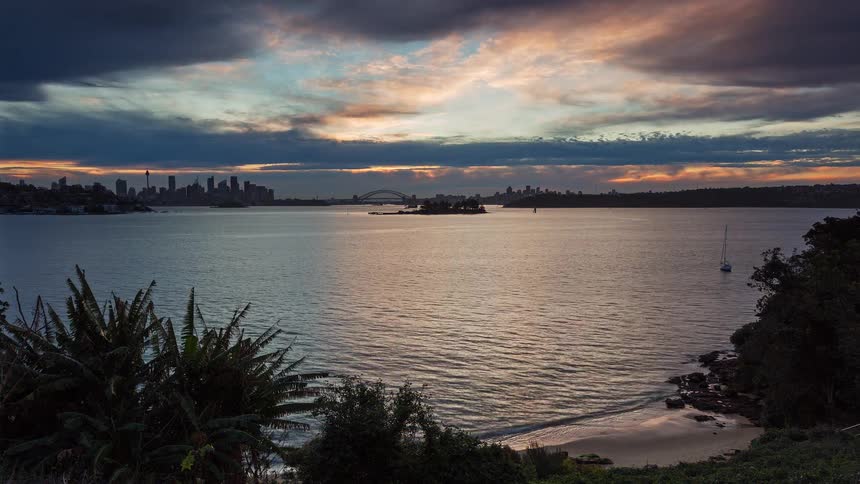 A timelapse shows the sun setting behind the sydney harbour bridge and city skyline casting a golden reflection across the water toward a quiet beachas the sky transitions from bright orange to deep blue the city lights begin to twinkle
