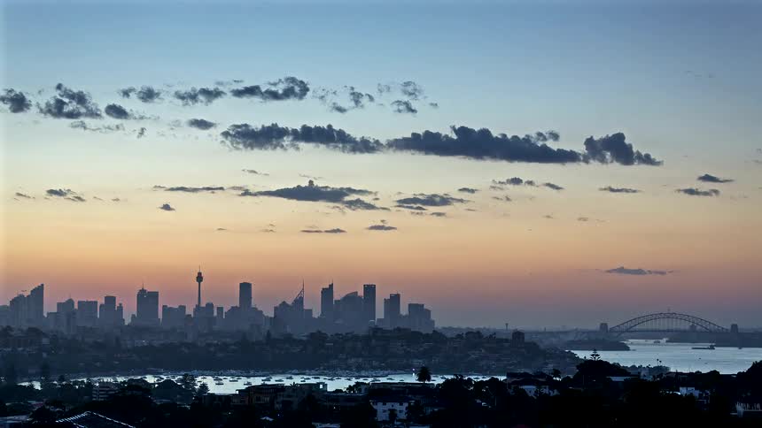 A time lapse view of a city skyline and harbor as the sky glows orange during sunset with clouds drifting above