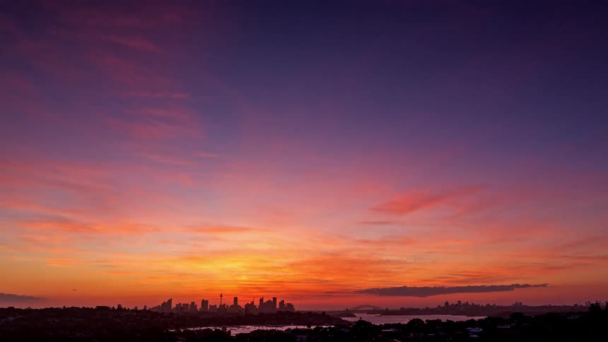 A distant city skyline glows under a vivid red and blue sunset sky as lights begin to appear across the landscape the features a river with boats