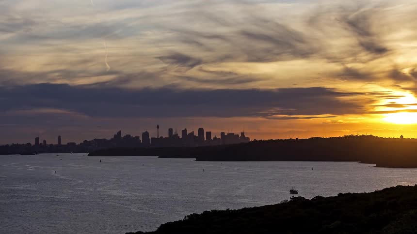 A time lapse view shows a distant city skyline across a wide harbor as the sun sets with streaks of orange and yellow light before turning into a dark night sky with illuminated buildings