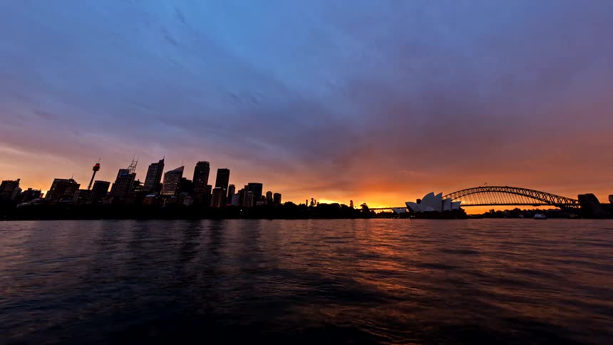 This timelapse captures a vibrant sunset over sydney harbour showcasing the city skyline opera house and harbour bridge as the sky transitions from soft blue to fiery orange the moving water reflects the shifting colors of the horizon wh