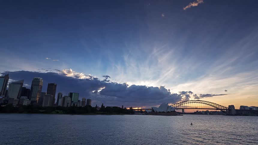 The sun sets behind the sydney opera house and harbour bridge as the city skyline begins to glow with interior lights under a deepening blue twilight sky filled with moving clouds over the dark harbor water