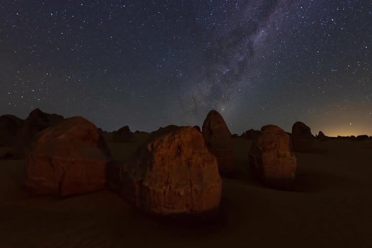 Rock formations stand in a desert under a starfilled night sky with the milky way stretching overhead