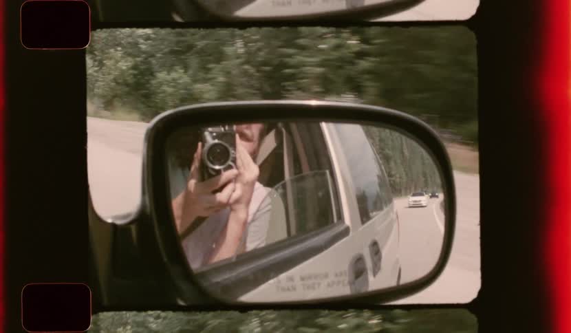 A person holds a vintage film camera while filming through the side view mirror of a moving car