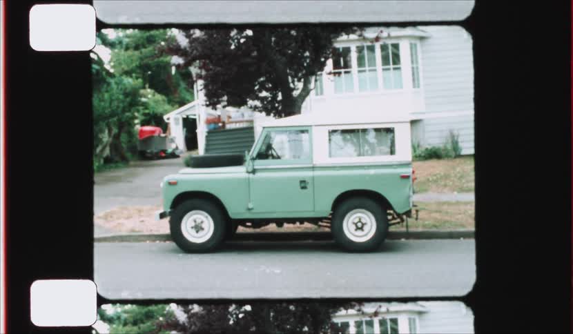 A green and white vintage off road vehicle is parked on a residential street in front of a house as seen through a retro film reel frame with visible sprocket holes and light grain