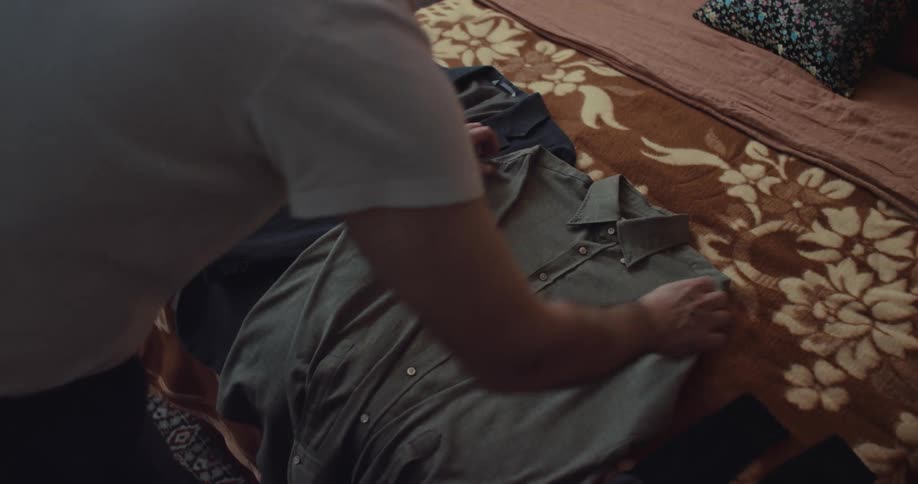 A man smooths out a grey button down shirt while organizing clothes on a bed