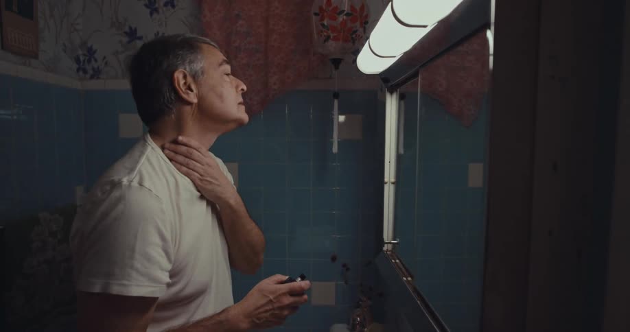 Man runs his hand over his neck while observing himself in the brightly lit bathroom mirror