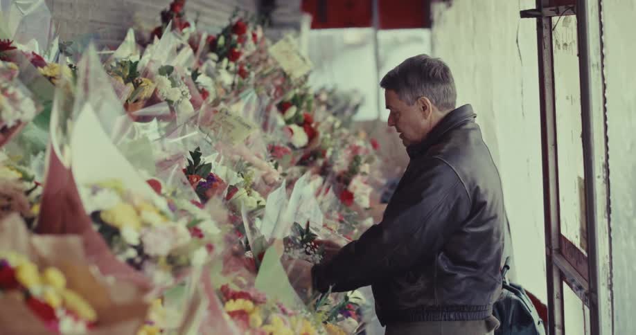A middle aged man wearing a dark leather jacket stands in a narrow outdoor market while carefully inspecting and choosing from rows of colorful flower bouquets wrapped in clear plastic