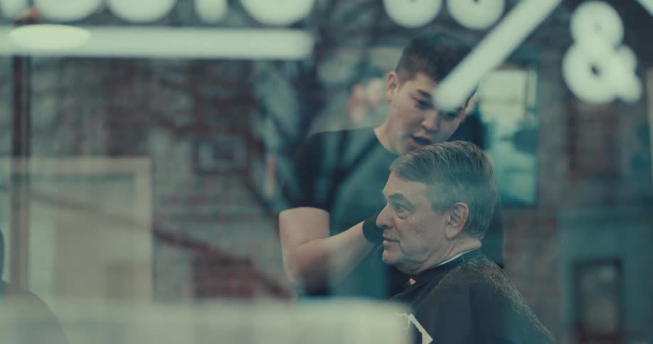 A barber wearing black gloves carefully trims the hair of a seated middle aged man inside a shop while they talk viewed through a window with a blurred foreground and brick walls in the background