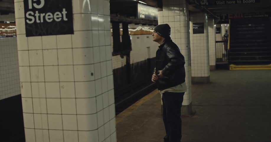 A man wearing a beanie and a black leather jacket stands alone on a subway platform marked 15 street looking down as he adjusts or reaches into his jacket while waiting beside the tiled columns and train tracks