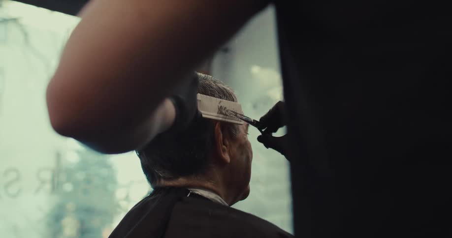 A barber wearing black gloves uses scissors and a comb to trim an elderly mans hair inside a barbershop