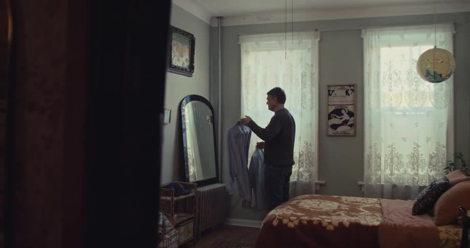 Man holds shirt in front of mirror inside bedroom lit by curtained windows