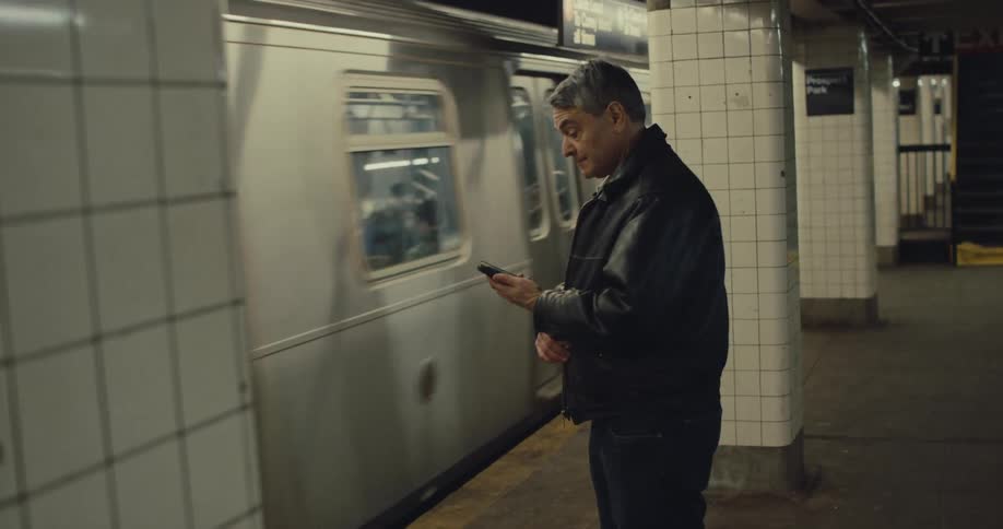A man in a black leather jacket stands on the subway platform and checks his smartphone as a silver train pulls into the station