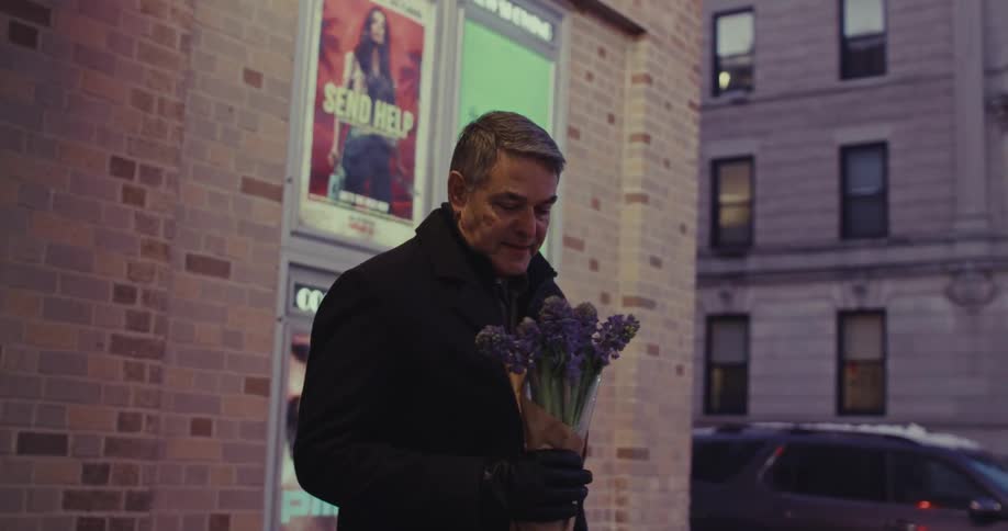A man in a dark coat stands outside a brick building at dusk is holding a small bouquet of purple flowers wrapped flowers while gazing ahead thoughtfully as city lights begin to glow around him
