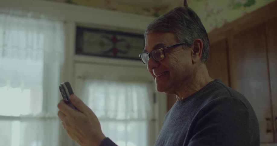 An older man with glasses stands in a kitchen and smiles warmly while looking at his smartphone screen during a video call