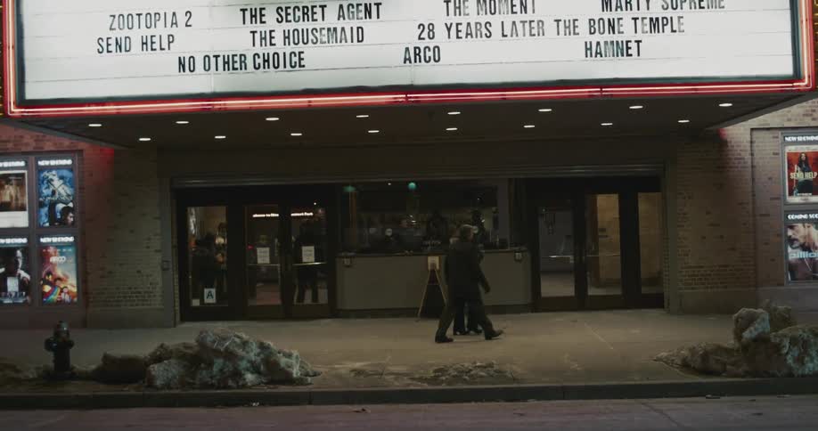 Two people stand talking outside and hug before entering a movie theater at night beneath a brightly lit marquee displaying film titles